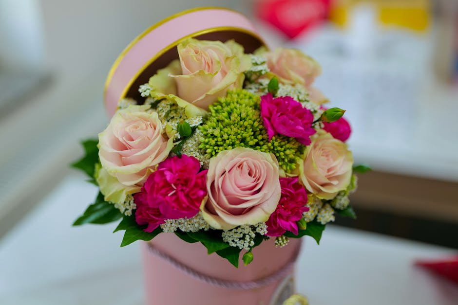 A round bouquet of fresh pale pink roses, vibrant pink carnations, and small white filler flowers arranged with green foliage, placed inside a pink cylindrical gift box with a matching ribbon. The bou