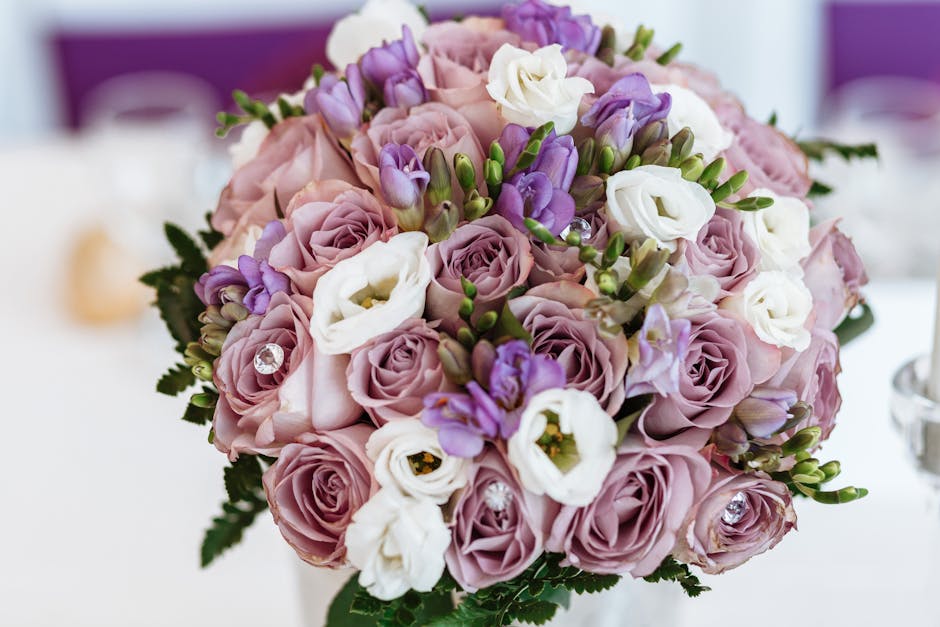 A round bouquet arrangement featuring soft pink roses, white lisianthus, and purple freesia, accented with small green foliage and subtle decorative crystals. The flowers are fresh, with layered petal