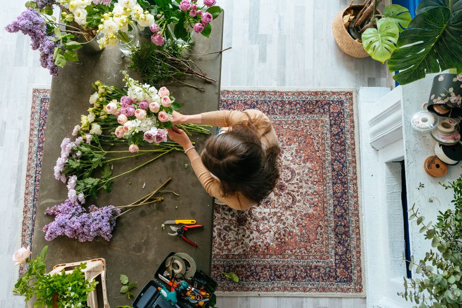 A florist with brown, shoulder-length hair arranging a bouquet of pink and white roses, hydrangeas, and other mixed flowers on a grey worktable indoors. The bouquet is partially assembled, with additi
